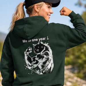 Könnte beinhalten: Ein dunkelgrüner Kapuzenpullover mit einem weißen Grafikdruck auf der Rückseite, der eine muskulöse Katze beim Anspannen ihres Arms zeigt. Über der Katze steht der Text "Me in one year ↓". Der Hoodie hat lange Ärmel und eine Kapuze.
