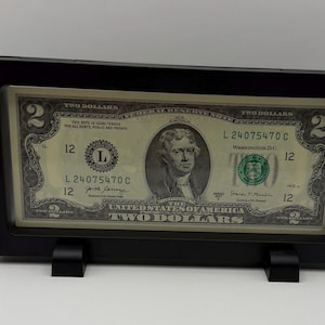 Könnte beinhalten: Ein gerahmter Zwei-Dollar-Schein in einem schwarzen Rahmen mit Ständer. Der Schein zeigt ein Porträt von Thomas Jefferson und den Text "TWO DOLLARS". Der Rahmen ist rechteckig und schlicht gestaltet.