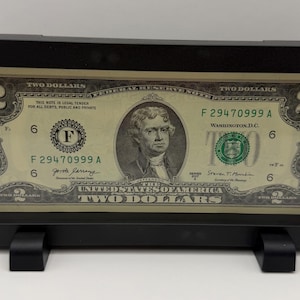 Op de afbeelding: Een ingelijste twee dollar biljet. De biljet toont een portret van Thomas Jefferson en de tekst "TWO DOLLARS". De zwarte lijst heeft een standaard.