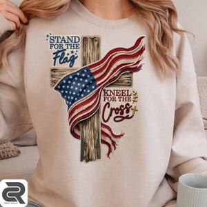 May include: A beige crewneck sweatshirt featuring a graphic print of a wooden cross draped with an American flag. The text reads "STAND FOR THE Flag" in blue and "KNEEL FOR THE Cross" in maroon, with small gold accents and white pearl-like trim.