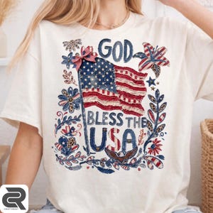 May include: A cream-colored t-shirt featuring a graphic print of an American flag, a red and white striped bow, and the text "GOD BLESS THE USA". The design includes floral elements, leopard print accents, and a bald eagle illustration in blue and red.