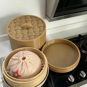 Könnte beinhalten: Ein hellbrauner, zylindrischer 3D-gedruckter Dampfgarer mit einem Deckel in Flechtoptik. In der offenen Basis liegt ein rundes, blassrosa Brötchen mit einer zentralen gefalteten Öffnung, einem kleinen roten Stern und zwei hellblauen Akzenten.