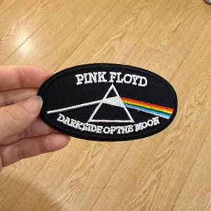 May include: An oval-shaped black embroidered patch featuring the Pink Floyd Dark Side of the Moon prism graphic. The design includes a white triangular prism, a white light beam, and a rainbow spectrum, with the text Pink Floyd Dark Side of the Moon.