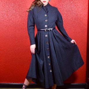 May include: A dark gray wool dress with a button-up front and a black belt. The dress has a full skirt and long sleeves. The model is wearing black heels and has a red background.