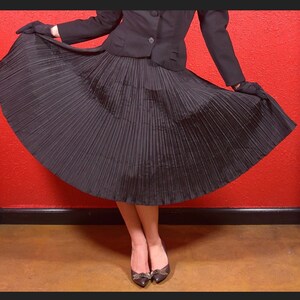 May include: A black pleated skirt with a fitted black jacket. The skirt is full and flowing, and the jacket has a classic, tailored look. The person is wearing black shoes with a bow.