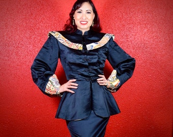 May include: A woman wearing a black satin jacket with a colorful patterned collar and cuffs. The jacket has a mandarin collar and a fitted silhouette. The woman is standing in front of a red wall.