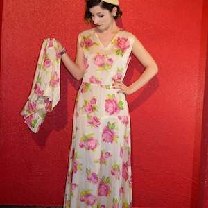 May include: A woman wearing a white dress with pink floral print and a white hat. She is standing in front of a red wall.