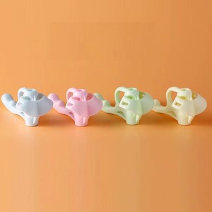 May include: Four plastic finger-positioning aids arranged in a row, featuring a light blue, a light pink, a light green, and a light yellow version. Each tool has a central ring for a finger and a curved extension for grip support.