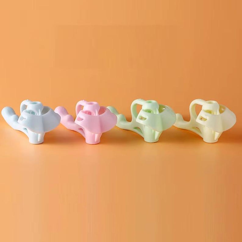 May include: Four plastic finger-positioning aids arranged in a row, featuring a light blue, a light pink, a light green, and a light yellow version. Each tool has a central ring for a finger and a curved extension for grip support.