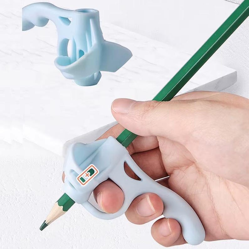 May include: A light blue ergonomic pencil grip featuring a trigger-like handle and a small rectangular label with a green plus sign icon. The grip is attached to a green hexagonal pencil, designed to assist with finger placement while writing.