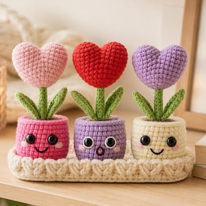 May include: Three crocheted heart-shaped flowers in pots, each with a unique face, sitting on a cream-coloured crocheted tray. The left pot is pink, the middle is purple, and the right is cream. Each flower has green leaves and a stem.