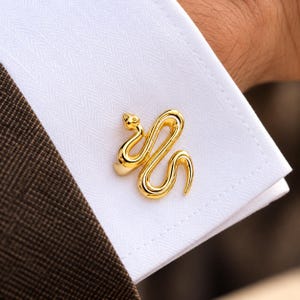Peut inclure: Un bouton de manchette en métal doré en forme de serpent enroulé avec une finition lisse et polie. L'accessoire est fixé sur le tissu blanc à texture chevron d'un poignet de chemise, mettant en valeur le design incurvé du corps du serpent.