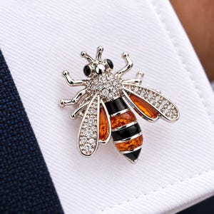 Peut inclure: Un bouton de manchette en forme d'abeille argentée avec un thorax incrusté de cristaux transparents, des rayures en émail noir et ambre sur l'abdomen, et des ailes ornées de cristaux et d'accents ambrés, sur un fond en tissu blanc.