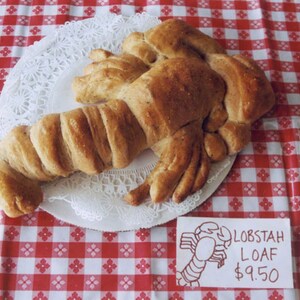 May include: A brown, baked good shaped like a lobster on a white doily on a red and white checkered tablecloth. The sign reads "Lobstah Loaf $9.50".