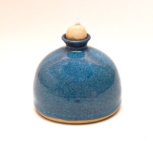 May include: A blue ceramic oil lamp with a round body and a small, round, light brown lid. The lamp has a white wick.