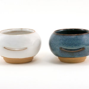 May include: Two ceramic bowls with a smiling face design. One bowl is white with a brown rim and the other is blue with a brown rim.