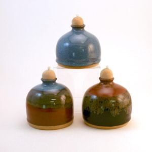 May include: Three ceramic oil lamps in shades of blue, brown, and green. Each lamp has a rounded body, a wooden stopper, and a wick. The lamps are displayed on a white surface, showcasing their unique colors and textures.