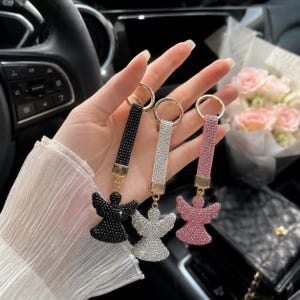 May include: Three angel-shaped keychains featuring a strap covered in small crystals and a matching crystal-encrusted angel pendant. The set includes one black, one silver, and one pink keychain, each attached to a gold-toned metal ring.