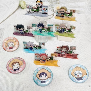 Op de afbeelding: Een verzameling acrylfiguurtjes met diverse anime-stijl personages in café-settings, samen met vier ronde buttons met afbeeldingen van personages die visvormige hoeden dragen, gerangschikt op een wit oppervlak met textuur.
