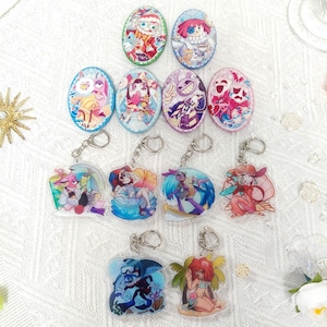 Könnte beinhalten: Eine Sammlung von zehn ovalen Anstecknadeln und Acryl-Schlüsselanhängern mit farbenfrohen Cartoon-Illustrationen. Die Designs zeigen verschiedene Figuren in lebhaften Outfits, einige mit Masken, Hüten oder strandbezogenen Accessoires wie Surfbrettern.