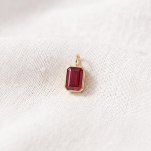 May include: A rectangular pendant featuring a deep red gemstone set in a polished gold-toned metal frame. The pendant includes a small, circular gold-toned bail at the top for attachment, showcasing a clean, geometric design with faceted edges.