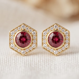 Puede incluir: Un par de pendientes de botón de oro, cada uno con una piedra preciosa central de color rojo intenso de corte redondo rodeada por un halo hexagonal de pequeñas piedras blancas claras y brillantes. El engaste es de oro pulido y brillante.