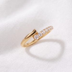 May include: A gold bypass ring featuring a polished band that tapers into a single end. One side of the band is set with five small, round, clear, faceted stones arranged in a row, creating a sparkling accent against the smooth gold metal finish.