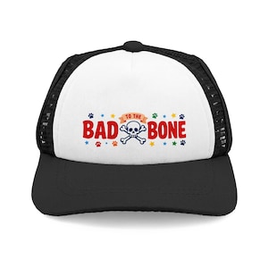 May include: A black and white trucker cap featuring a graphic on the front panel with the red text BAD BONE, the orange text TO THE, and a white skull and crossbones icon. The design is surrounded by small colourful paw prints and stars.