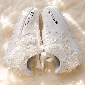 Puede incluir: Un par de zapatillas blancas con aplicaciones florales de encaje blanco, cordones de cinta de satén y pequeños detalles de perlas. El zapato izquierdo muestra la fecha 24/10/09 y el derecho tiene el texto Mrs. Eyre impreso en negro.