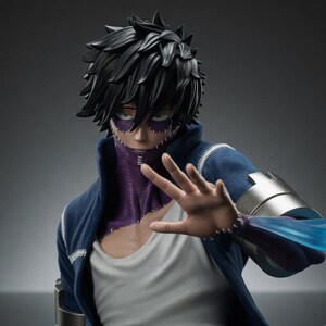 Puede incluir: Figura detallada de un personaje con cabello negro puntiagudo, cicatrices faciales moradas y una chaqueta azul. La figura posa con la mano extendida y un efecto de energ&iacute;a azul emana del brazo.
