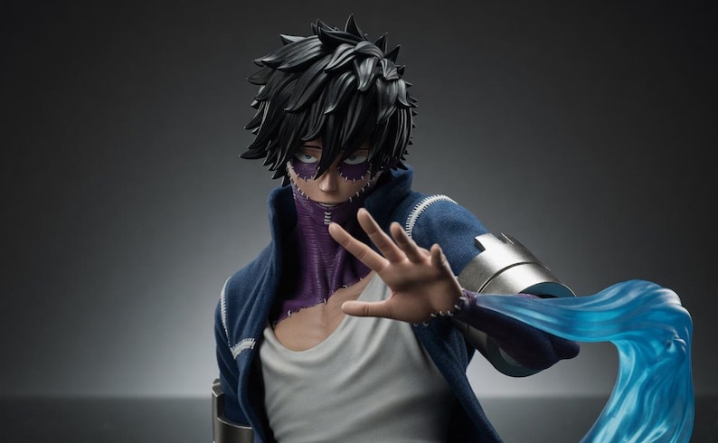Puede incluir: Figura detallada de un personaje con cabello negro puntiagudo, cicatrices faciales moradas y una chaqueta azul. La figura posa con la mano extendida y un efecto de energ&iacute;a azul emana del brazo.