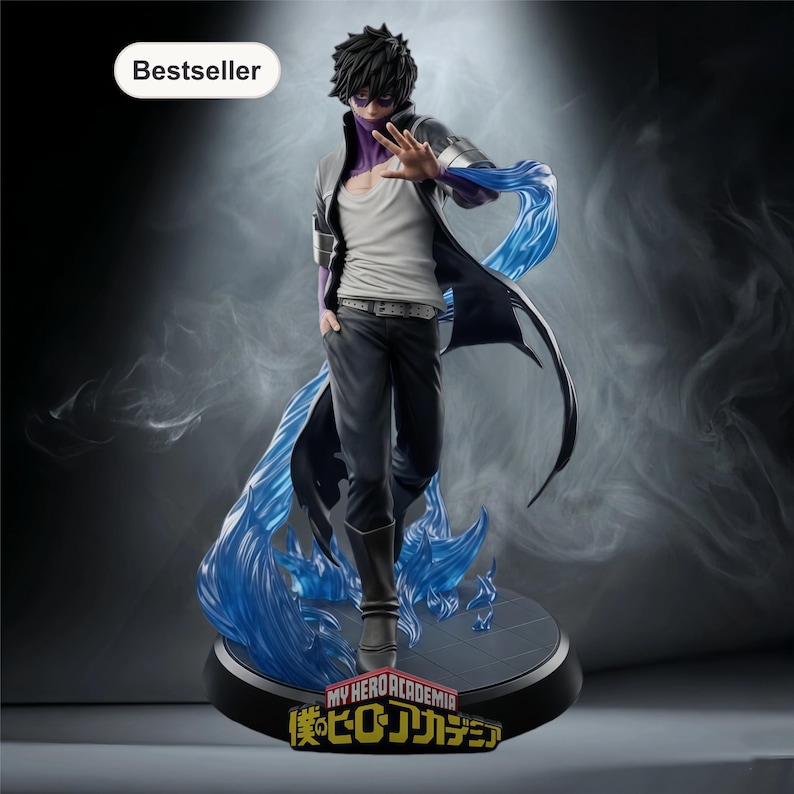 Puede incluir: Figura detallada de un personaje de "My Hero Academia" con cabello negro y marcas moradas en la piel. La figura lleva una camisa blanca, pantalones negros y un abrigo largo, con llamas azules que emanan de la base. La base tiene el logo del programa y la palabra "Bestseller".