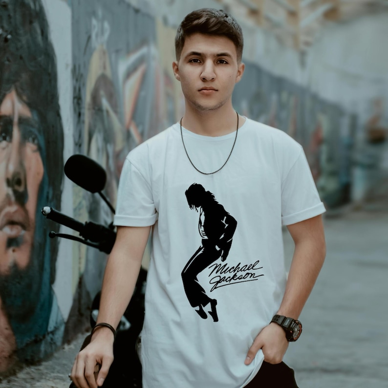 May include: A white short-sleeved crew neck t-shirt featuring a black graphic silhouette of Michael Jackson in a dance pose, accompanied by the signature Michael Jackson text logo printed in black on the front centre of the garment.