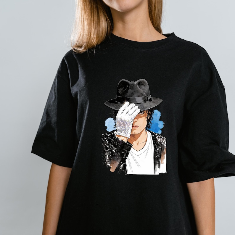 May include: A black t-shirt featuring a graphic illustration of a person wearing a black fedora and a white sequined glove covering their eyes. The figure wears a black sequined jacket over a white v-neck shirt, set against a blue watercolour splash.