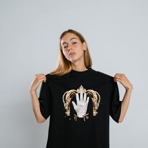 May include: A black t-shirt featuring a graphic print of a gold crown framing a single white sequined glove. The glove is positioned centrally within the crown, which has intricate gold detailing and clear crystal accents on a dark fabric.