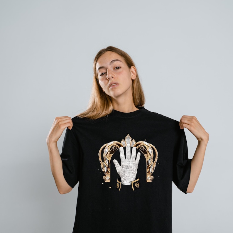 May include: A black t-shirt featuring a graphic print of a gold crown framing a single white sequined glove. The glove is positioned centrally within the crown, which has intricate gold detailing and clear crystal accents on a dark fabric.