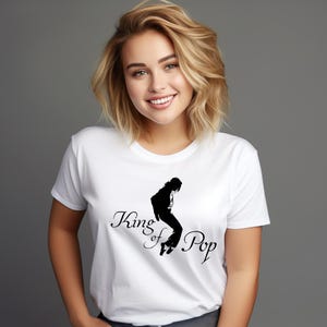 May include: A white short-sleeved t-shirt featuring a black graphic silhouette of a person in a dancing pose. The text "King of Pop" is printed in a black cursive script font across the centre of the chest, positioned below the silhouette.