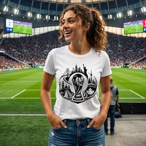 May include: A white short-sleeved t-shirt featuring a black graphic print of a soccer trophy centered above the text 2026, surrounded by silhouettes of iconic landmarks including a bridge, a tower, and a maple leaf, worn with blue denim jeans.