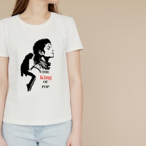 May include: A white short-sleeved t-shirt featuring a black graphic of a person in profile and a silhouette of a person dancing. The text "THE" and "OF POP" are printed in black, while the word "King" is printed in red below the profile.