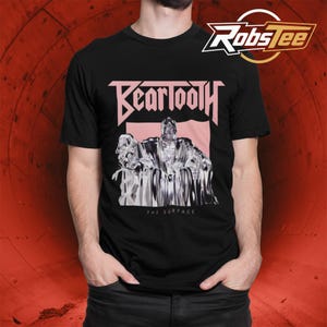 Puede incluir: Una camiseta negra de manga corta con la palabra Beartooth en letras rosa claro sobre un gráfico de una figura metálica sentada en un trono. Debajo del gráfico, el texto dice The Surface. La camiseta tiene cuello redondo y corte estándar.