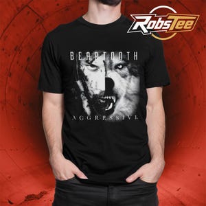 May include: A black short-sleeve t-shirt featuring a graphic print of a human face merged with a wolf face in black and white. The text "BEARTOOTH" appears above the graphic, and the word "AGGRESSIVE" is printed below the graphic in white.