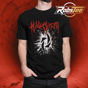May include: Black t-shirt featuring the band name "Halestorm" in red, dripping font above a white and black graphic. The graphic is a stylized design with two arrows. The shirt has a crew neck.