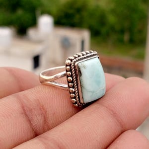 May include: A silver ring featuring a rectangular, light blue stone with a smooth surface. The setting is framed by a decorative border of small metal beads and a twisted rope pattern. The band is split into two thin, polished silver segments.