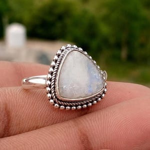 May include: A silver ring featuring a triangular moonstone cabochon set in a bezel. The stone is surrounded by a decorative border of small silver beads and a twisted rope texture. The band is smooth and polished, made of sterling silver.