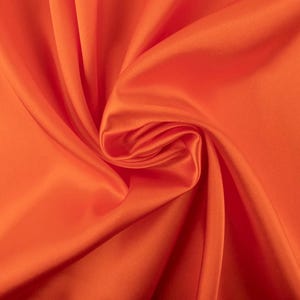 May include: A vibrant orange fabric with a smooth, lustrous finish. The material is draped in soft, flowing folds that create deep shadows and bright highlights across the surface, emphasizing the consistent, solid color and silky texture.