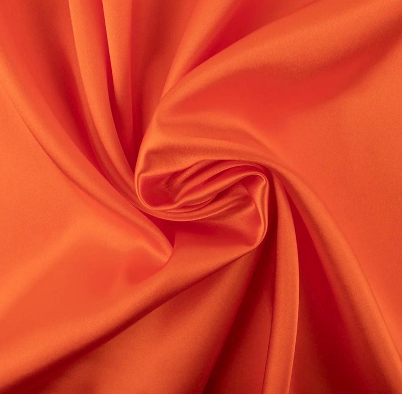 May include: A vibrant orange fabric with a smooth, lustrous finish. The material is draped in soft, flowing folds that create deep shadows and bright highlights across the surface, emphasizing the consistent, solid color and silky texture.