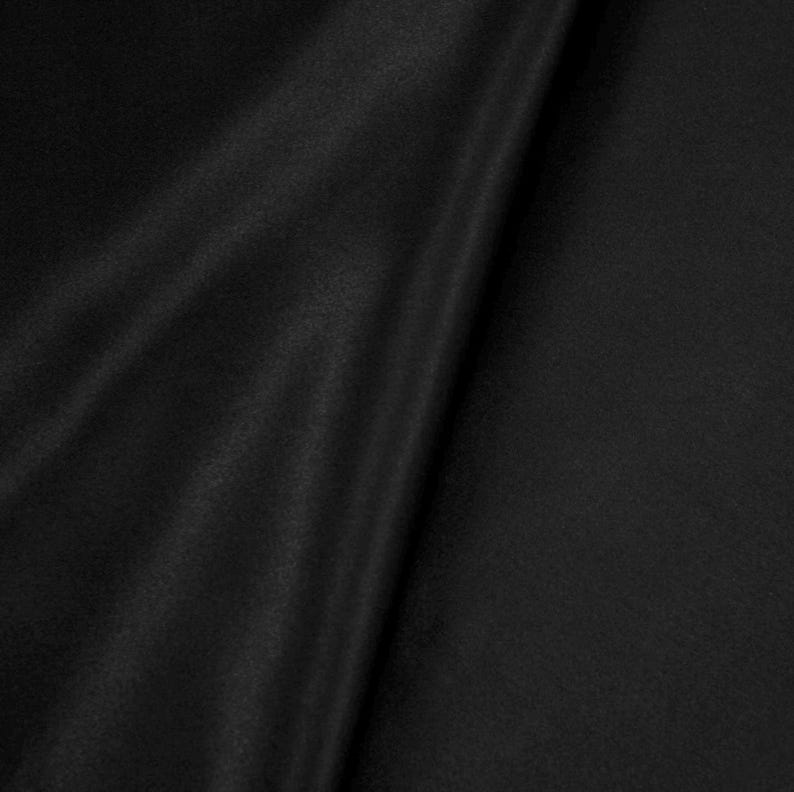 May include: A solid black fabric with a smooth, slightly reflective surface texture. The material is draped to create soft, diagonal folds that catch the light, highlighting the deep, uniform black color and the fine, consistent weave of the textile.