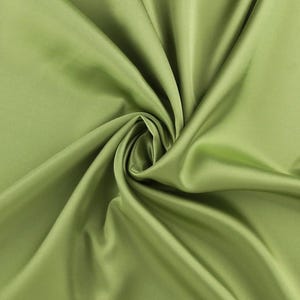 May include: A solid light olive green fabric with a smooth, lustrous finish. The material is draped in a central spiral, creating deep, flowing folds and shadows that highlight the reflective quality and uniform color of the textile surface.