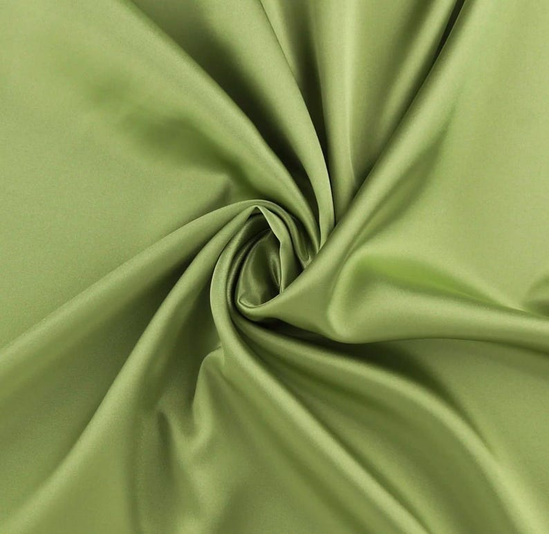 May include: A solid light olive green fabric with a smooth, lustrous finish. The material is draped in a central spiral, creating deep, flowing folds and shadows that highlight the reflective quality and uniform color of the textile surface.