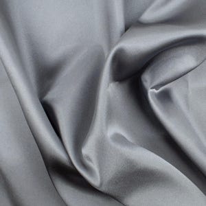 May include: A solid grey fabric with a smooth, lustrous finish. The material is draped in soft, flowing folds that create deep shadows and bright highlights across the surface, emphasizing the reflective quality and texture of the textile.